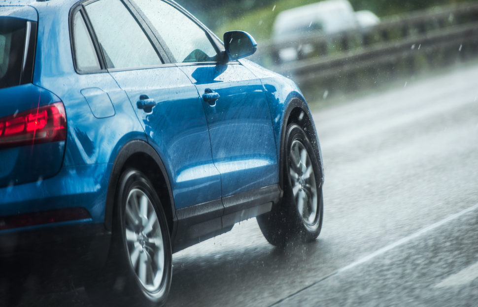 Coche azul conduciendo bajo la lluvia en carretera, resaltando la importancia de mantener las lunas del coche en invierno para una mejor visibilidad y seguridad.